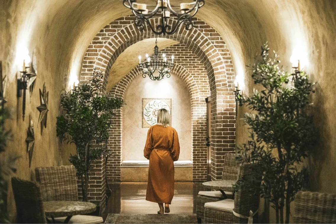 A woman in an orange robe walks down a beautifully arched brick hallway adorned with greenery and soft lighting.