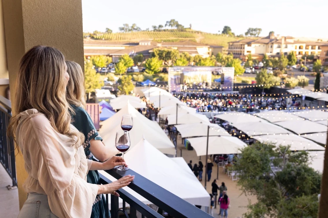 Two women enjoy wine on a balcony overlooking a festival with tents and a crowd, set against a backdrop of Napa Valley vineyards.