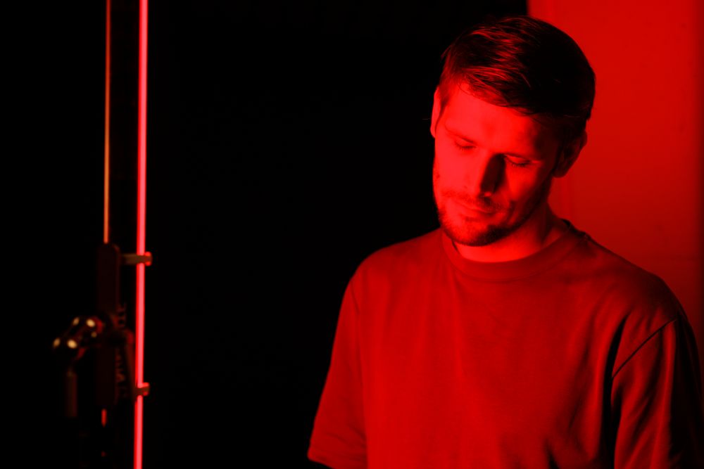 A man with short hair stands in a dimly lit space, illuminated by red LED light, appearing thoughtful with his eyes closed.