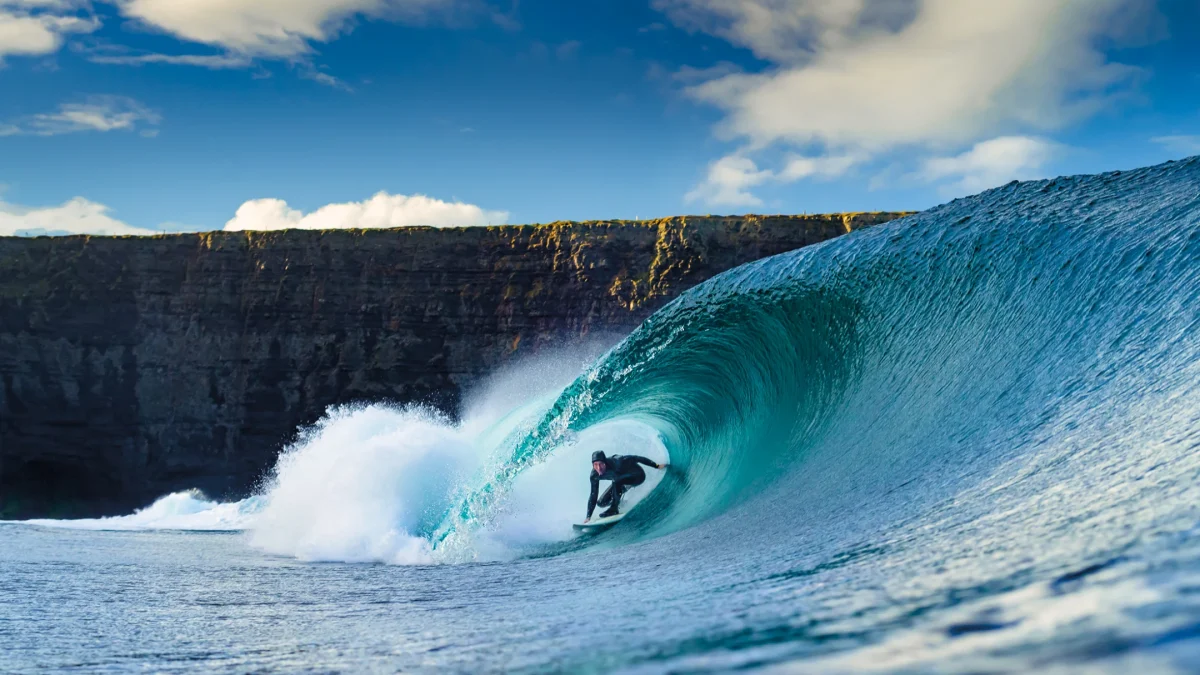 Surfer riding a large wave with a cliff backdrop under a blue sky and scattered clouds.