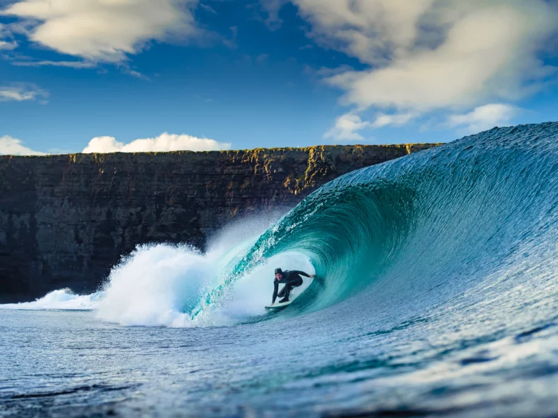 Surfer riding a large wave with a cliff backdrop under a blue sky and scattered clouds.
