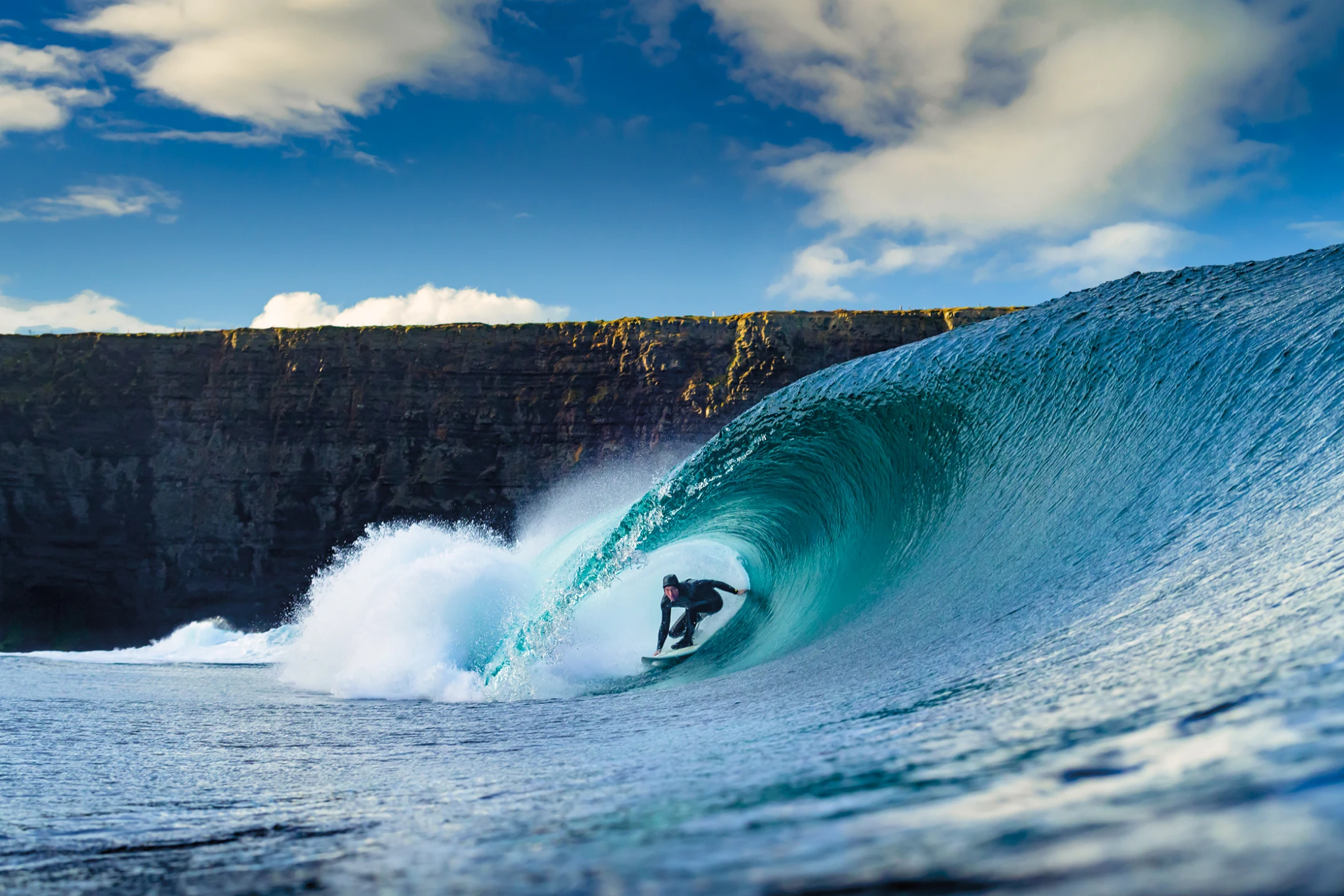 Surfer riding a large wave with a cliff backdrop under a blue sky and scattered clouds.