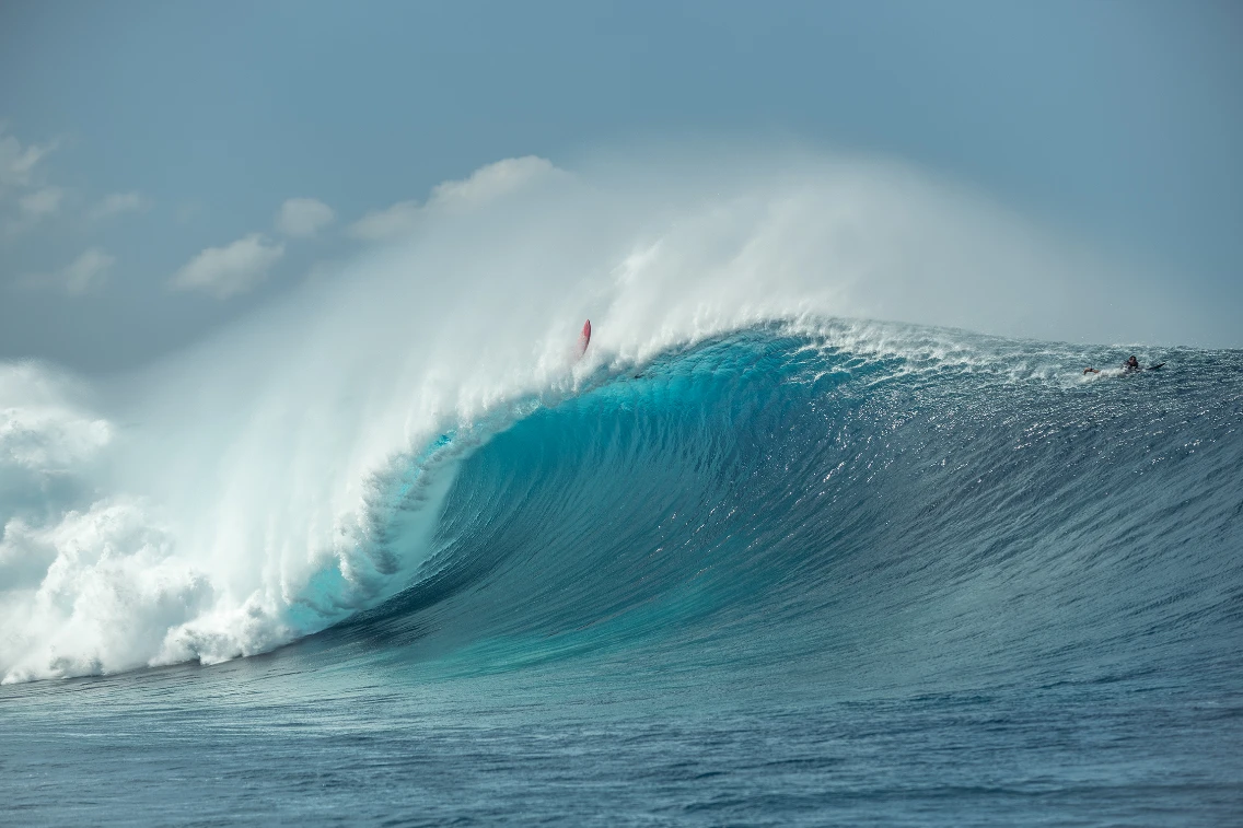 A large, powerful wave crashes in the ocean with a surfer riding the crest, showcasing big-wave surf conditions.