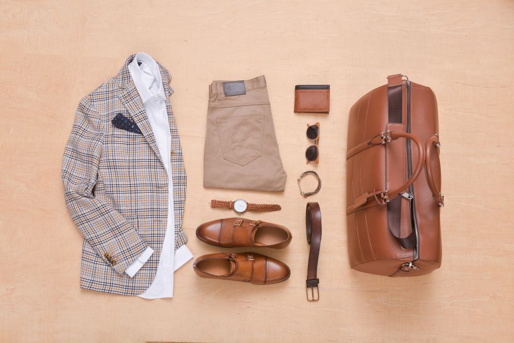 Men's luxury fashion laid out, featuring a plaid blazer, trousers, accessories, and a leather weekend bag for fall 2026 trends.