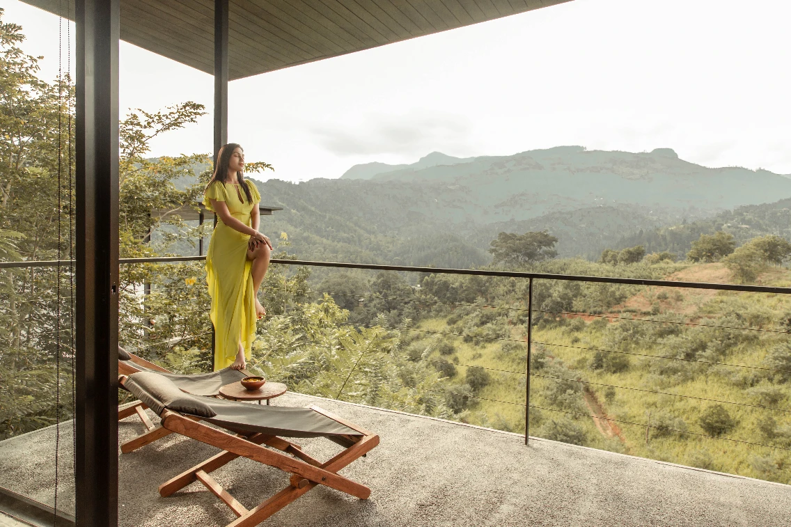 A woman in a yellow dress stands on a balcony, enjoying a scenic view of mountains and lush greenery in Sri Lanka.