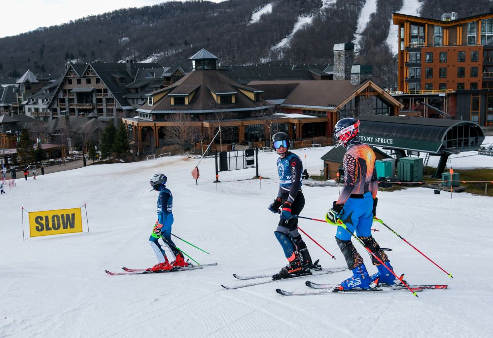 Skiers navigate a snowy slope at Spruce Peak with resort buildings in the background and a warning sign for slow skiing.