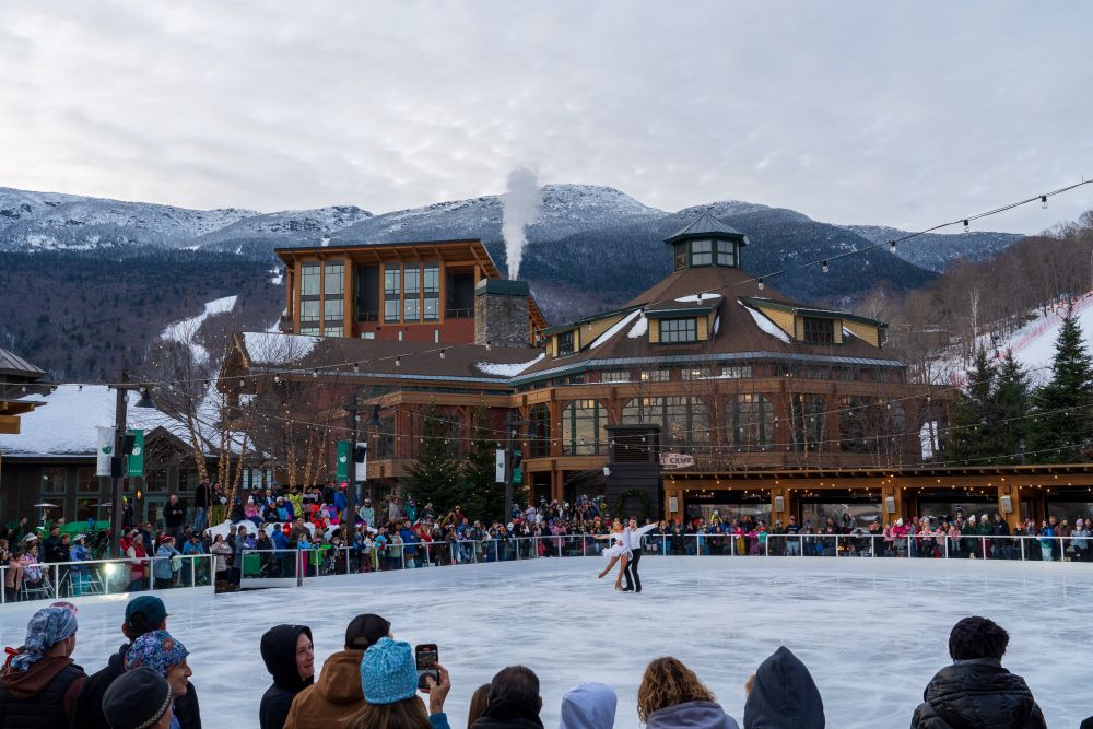 Skaters perform on an outdoor ice rink surrounded by spectators and the Lodge at Spruce Peak in the snowy mountains of Vermont.