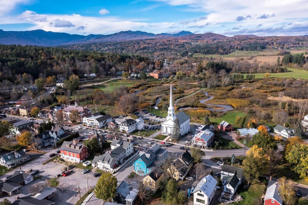 Aerial view of a picturesque New England town with colorful buildings, a church steeple, and surrounding mountains.