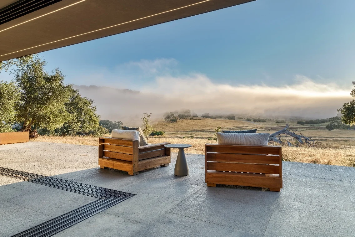 Seating area with wooden chairs and a table overlooking a misty valley and lush landscape from inside the Meadow House.