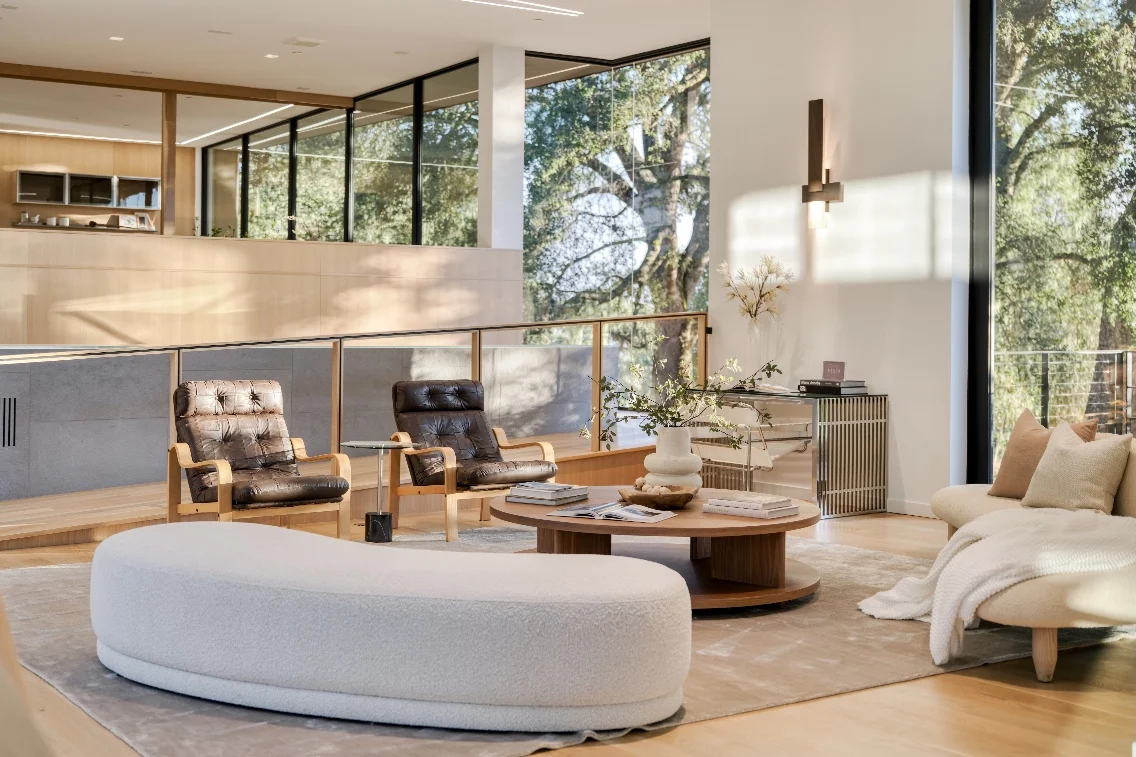 Modern living room featuring a curved white sofa, brown leather chairs, and large windows overlooking natural greenery.