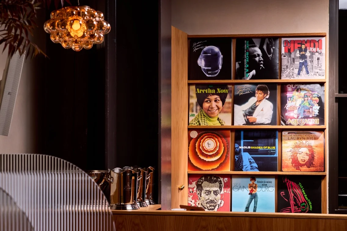 A display of vinyl records on a wooden shelf, featuring various iconic artists and a decorative light fixture above.
