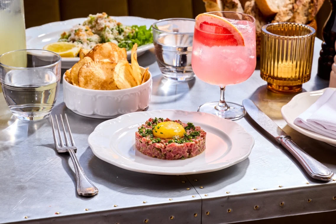 A plate of beef tartare topped with an egg yolk, accompanied by crispy potato chips and a pink cocktail on a dining table.