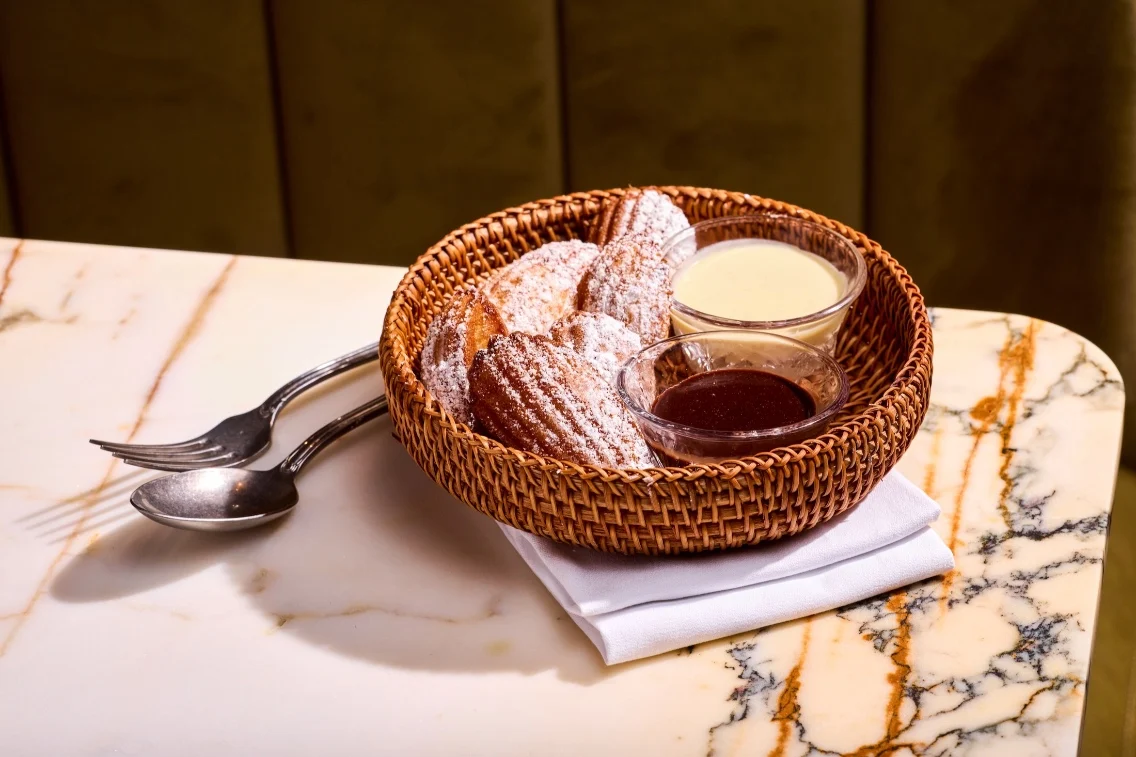 A basket of madeleines dusted with powdered sugar, accompanied by bowls of chocolate and vanilla sauces.