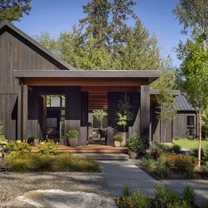 Contemporary black wood-paneled home entrance surrounded by lush greenery and a gravel pathway.