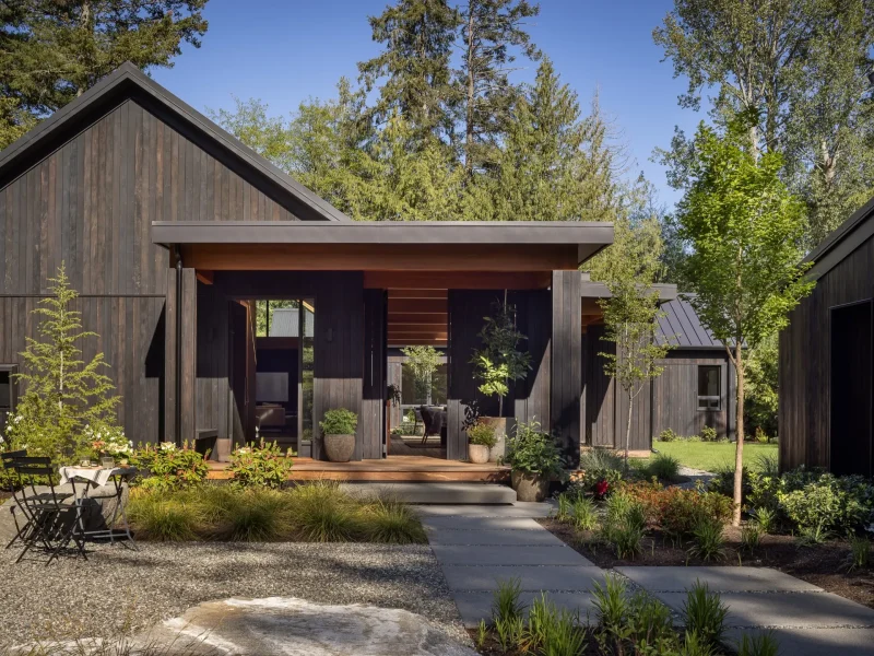 Contemporary black wood-paneled home entrance surrounded by lush greenery and a gravel pathway.