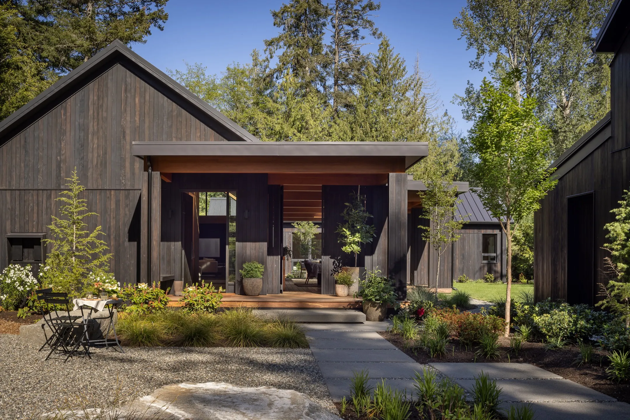 Contemporary black wood-paneled home entrance surrounded by lush greenery and a gravel pathway.