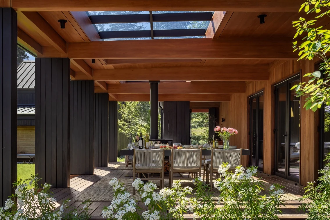 Modern outdoor dining area featuring natural wood beams, a table set up for a meal, and flowers in the foreground.