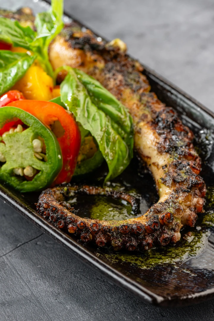Grilled octopus served with fresh herbs and colorful salad vegetables on a black plate.