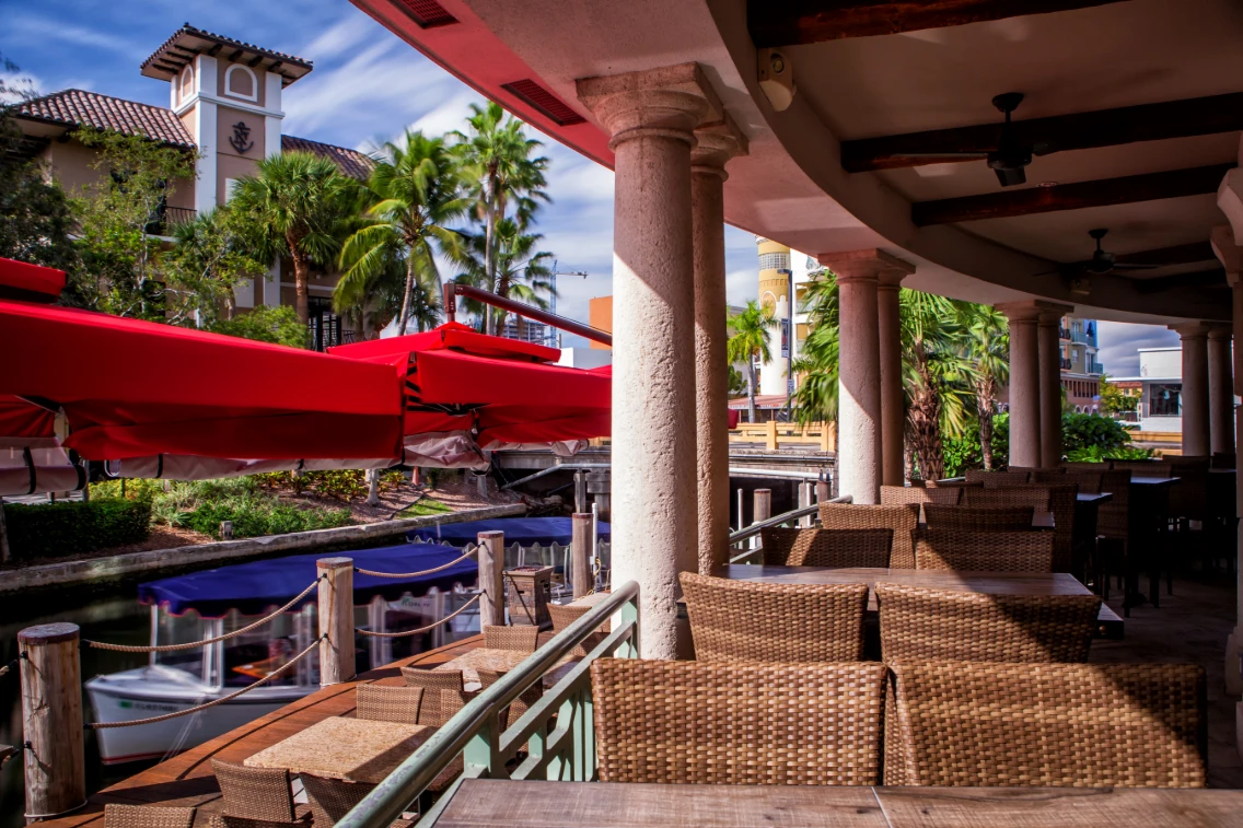 Outdoor dining area with wicker furniture, red umbrellas, and palm trees along the Las Olas waterfront at Casa Sensei.