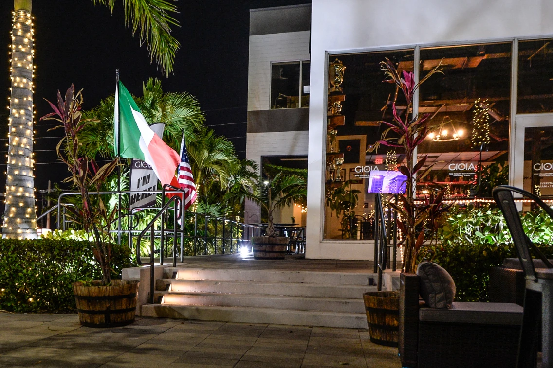 Entrance to Gioia Italy Restaurant featuring Italian and American flags, vibrant plants, and decorative lighting at night.
