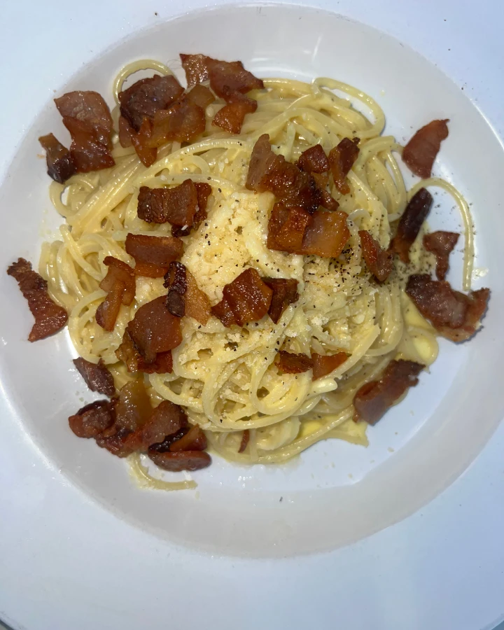 Spaghetti carbonara topped with crispy pancetta and grated cheese, served in a white dish.
