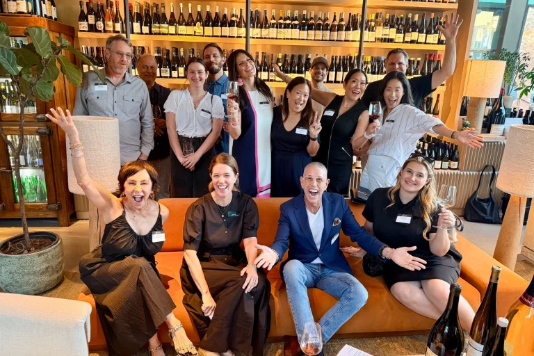 A joyful group of people gathered at a wine event, celebrating local cuisine while holding glasses and smiling.