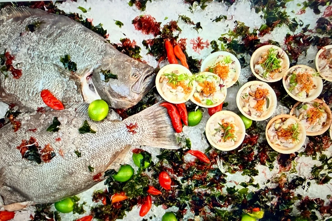 Fresh fish displayed on ice alongside bowls of seafood dishes garnished with herbs and limes, amid colorful garnishes and peppers.