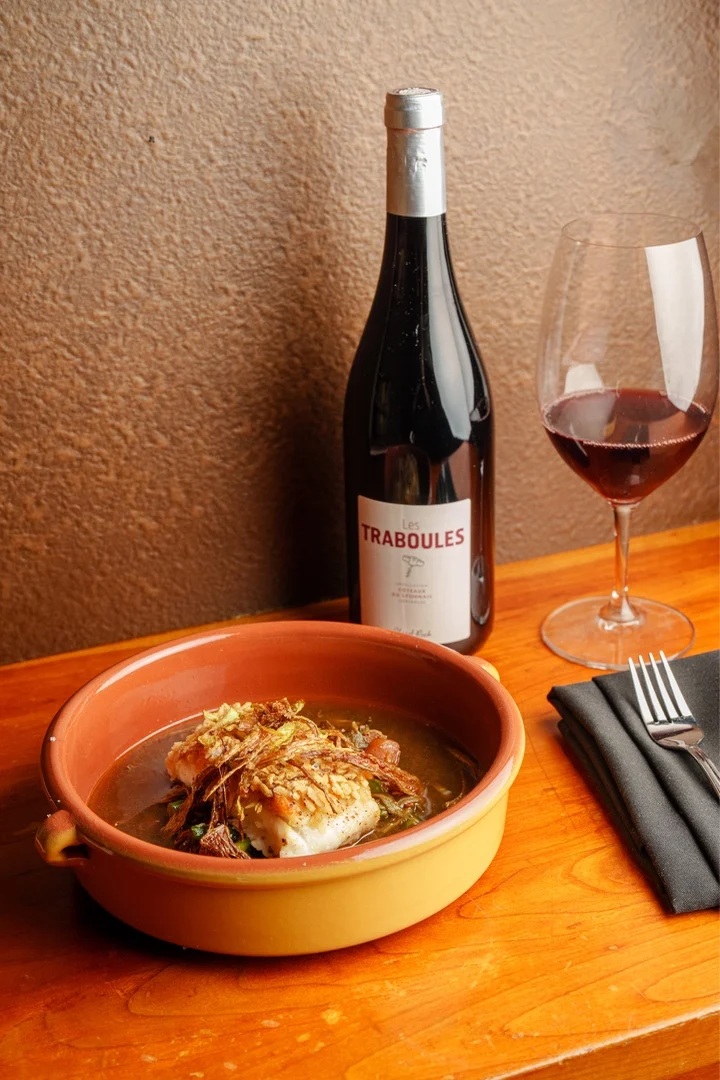 A bowl of seasoned fish garnished with crispy shallots, a wine bottle, and a glass of red wine on a wooden table.