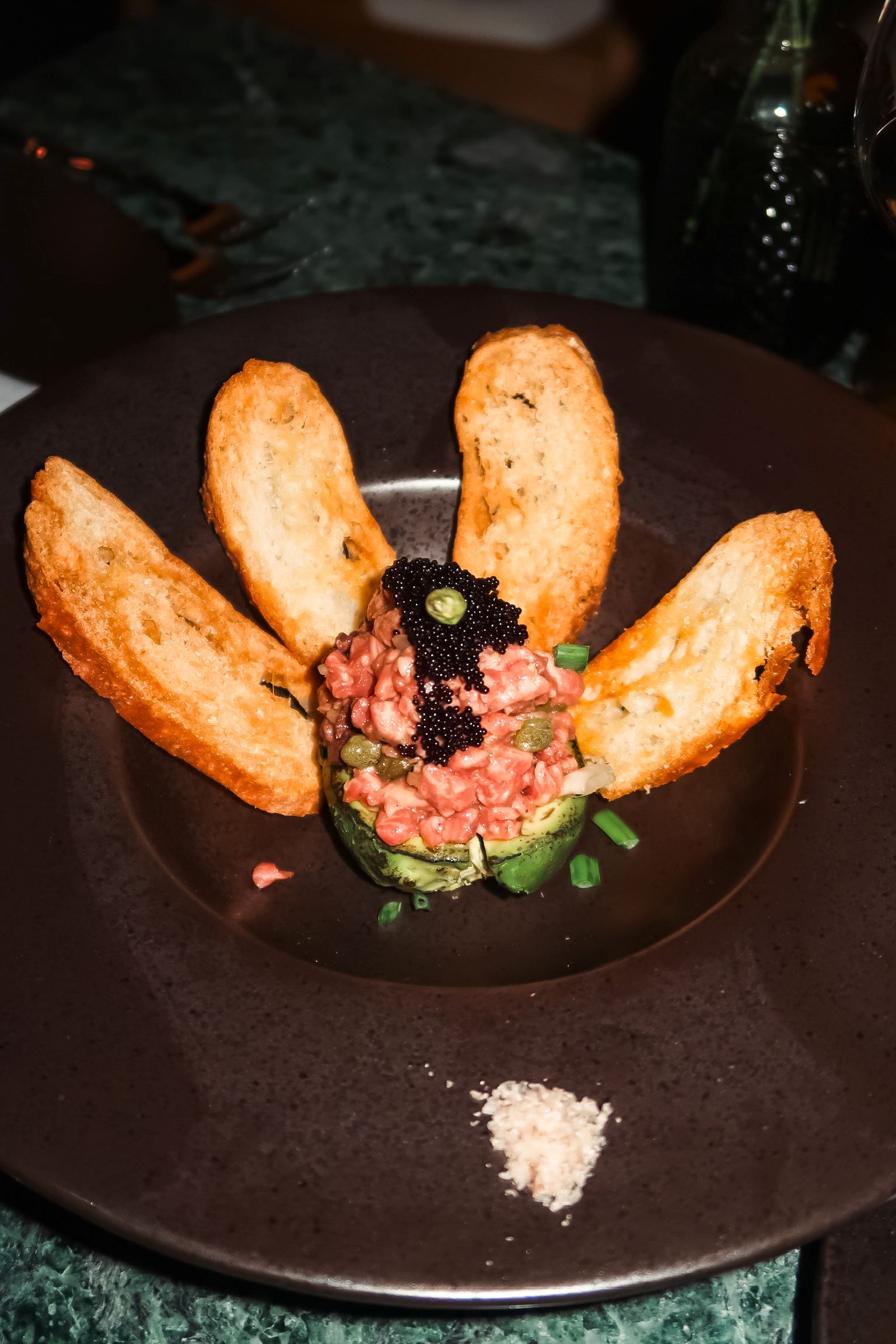 Wagyu tartare served with crispy bread slices and caviar on a dark plate, garnished with green onions and sea salt.