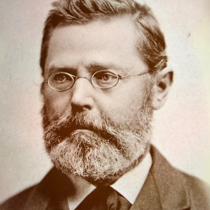 Black and white portrait of a man with glasses and a beard, dressed in a dark suit and tie.