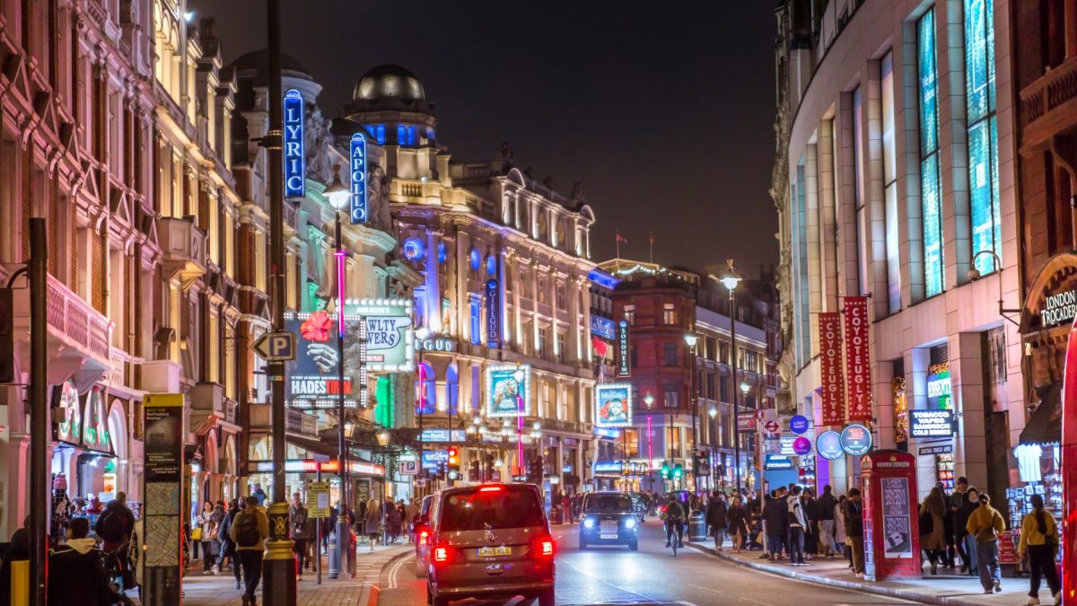 Busy city street at night with brightly lit theaters, cars, and pedestrians enjoying a lively atmosphere.