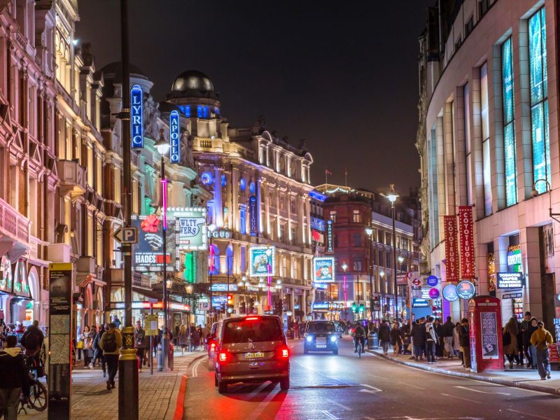 Busy city street at night with brightly lit theaters, cars, and pedestrians enjoying a lively atmosphere.
