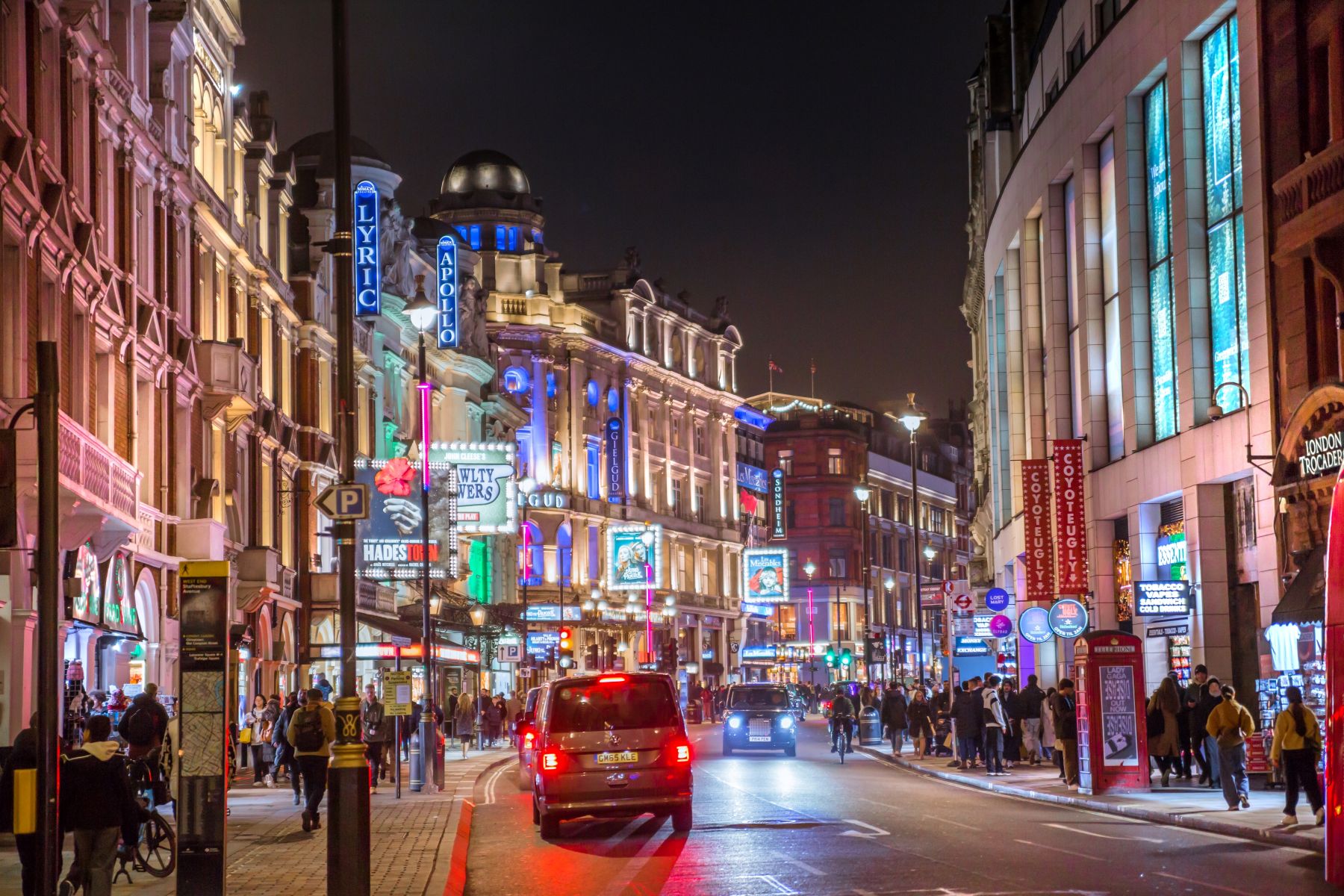 Busy city street at night with brightly lit theaters, cars, and pedestrians enjoying a lively atmosphere.