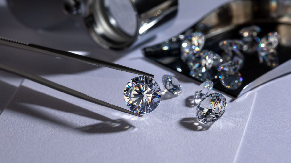 A shiny lab-grown diamond held by tweezers, surrounded by additional diamonds and a magnifying glass on a white surface.