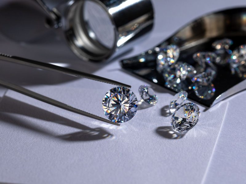 A shiny lab-grown diamond held by tweezers, surrounded by additional diamonds and a magnifying glass on a white surface.