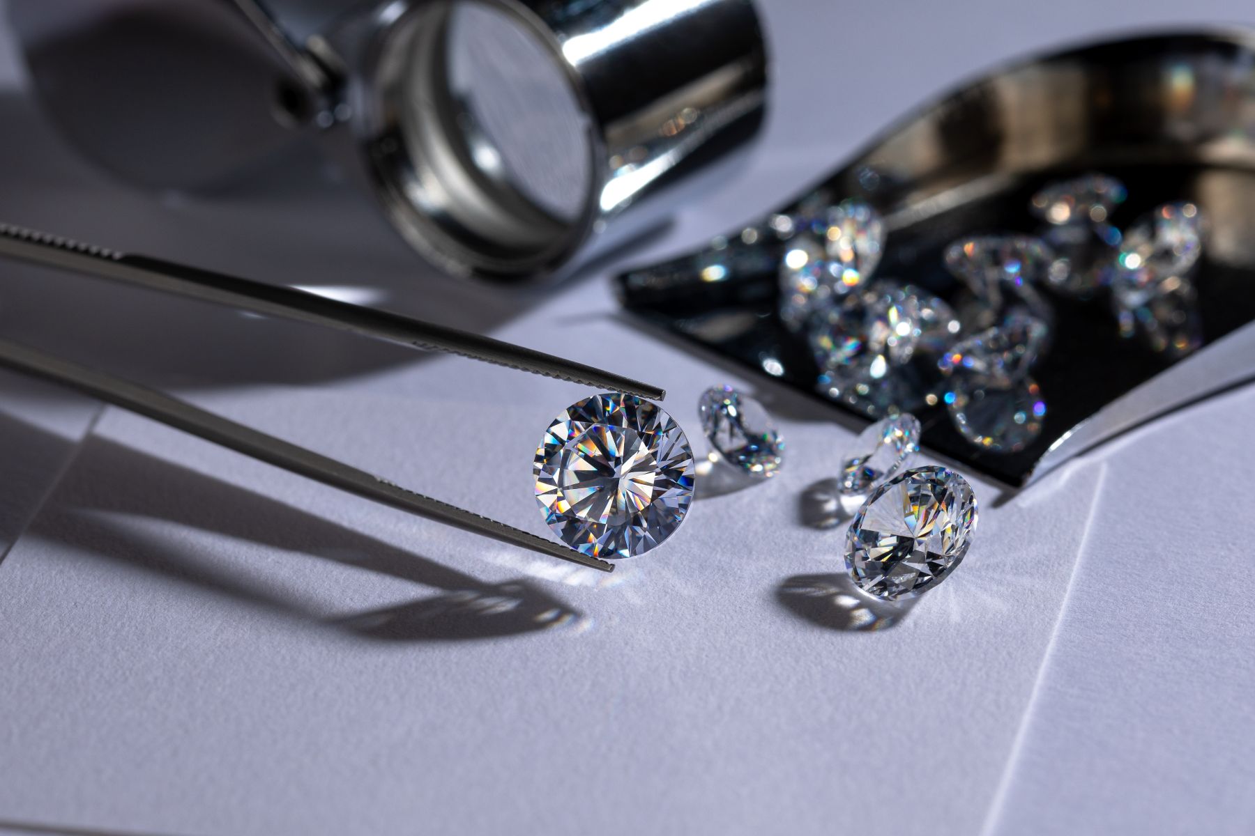 A shiny lab-grown diamond held by tweezers, surrounded by additional diamonds and a magnifying glass on a white surface.