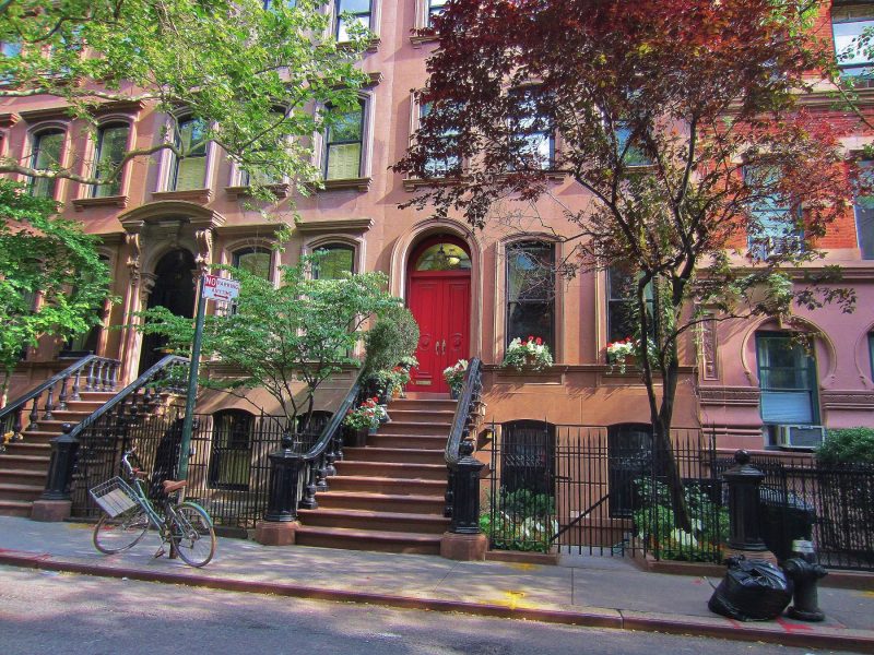 Brownstone building with a red door, surrounded by greenery and a bike parked outside on the Upper West Side of Manhattan.