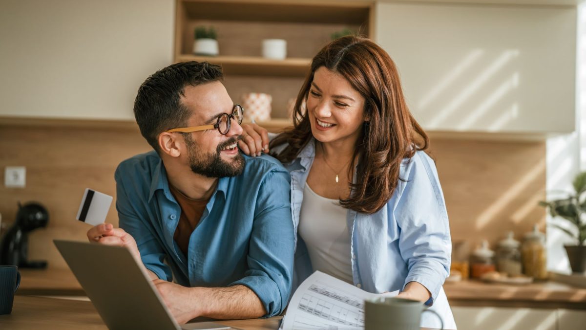 Couple discussing major lifestyle purchases at a kitchen table with a laptop, documents, and a credit card.