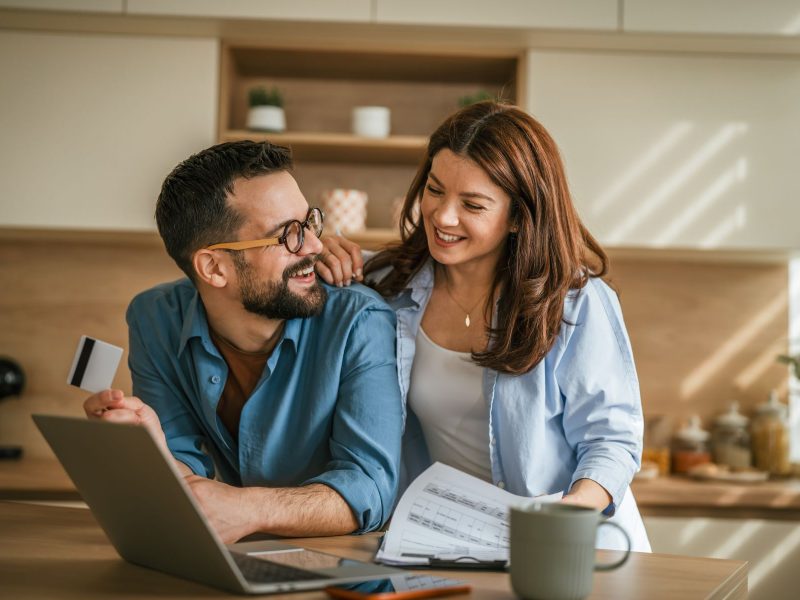 Couple discussing major lifestyle purchases at a kitchen table with a laptop, documents, and a credit card.