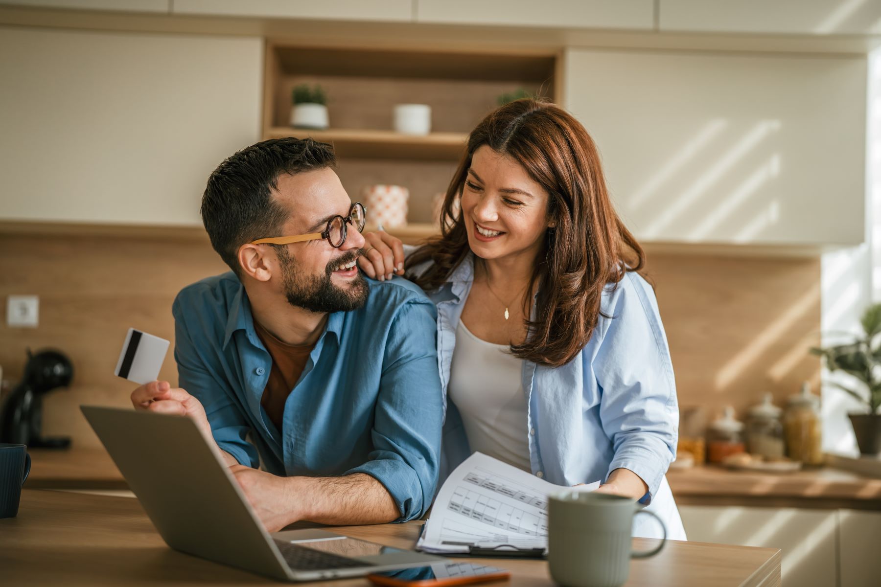 Couple discussing major lifestyle purchases at a kitchen table with a laptop, documents, and a credit card.