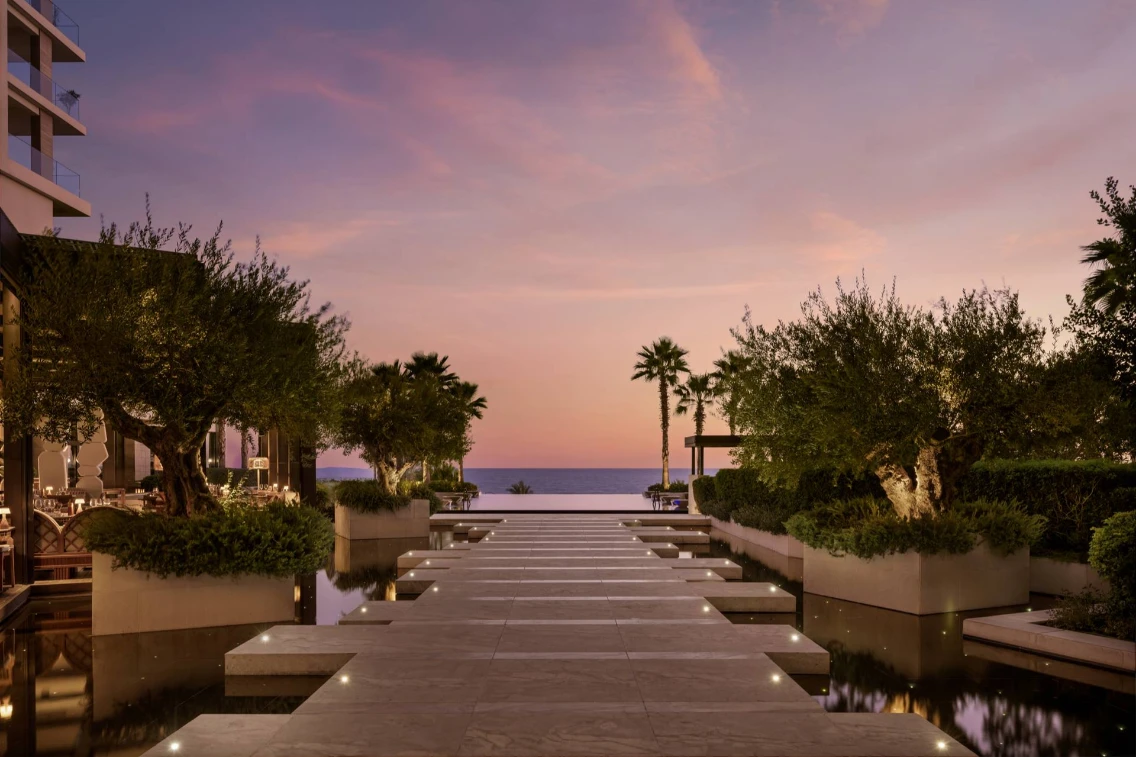 Elegant pathway lined with olive trees leads to the sea at the AMARA Hotel, showcasing a stunning sunset view.