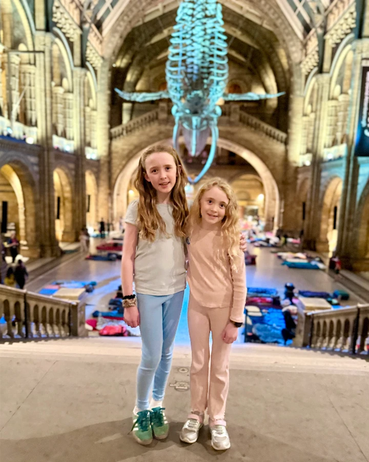 Two girls pose together in front of a whale skeleton at the Natural History Museum during a Kids' Dino Snores event.