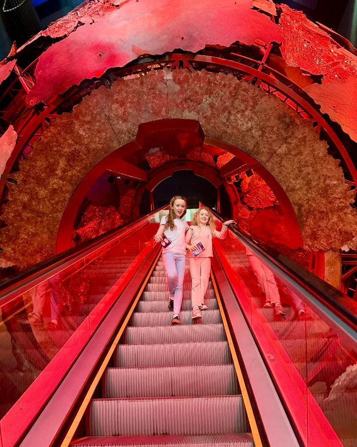 Two children descend an escalator, smiling and holding tickets, surrounded by vibrant red and earthy tones at the museum.