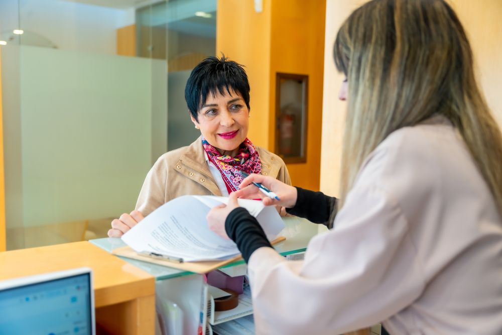 A woman with short dark hair smiles while speaking with a staff member at a medical-aesthetic reception desk.