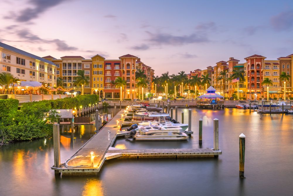 Marina with boats at sunset, surrounded by colorful buildings and palm trees in Naples, Florida's luxury waterfront area.