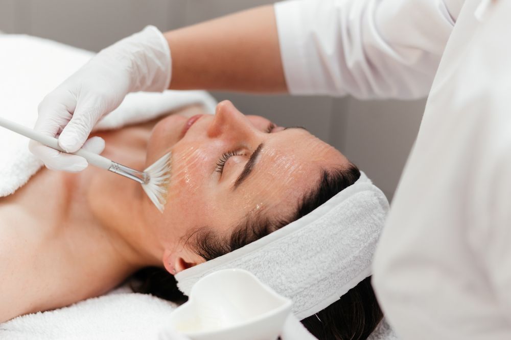 A woman receives a facial treatment with a brush in a spa setting, surrounded by towels and skincare products.