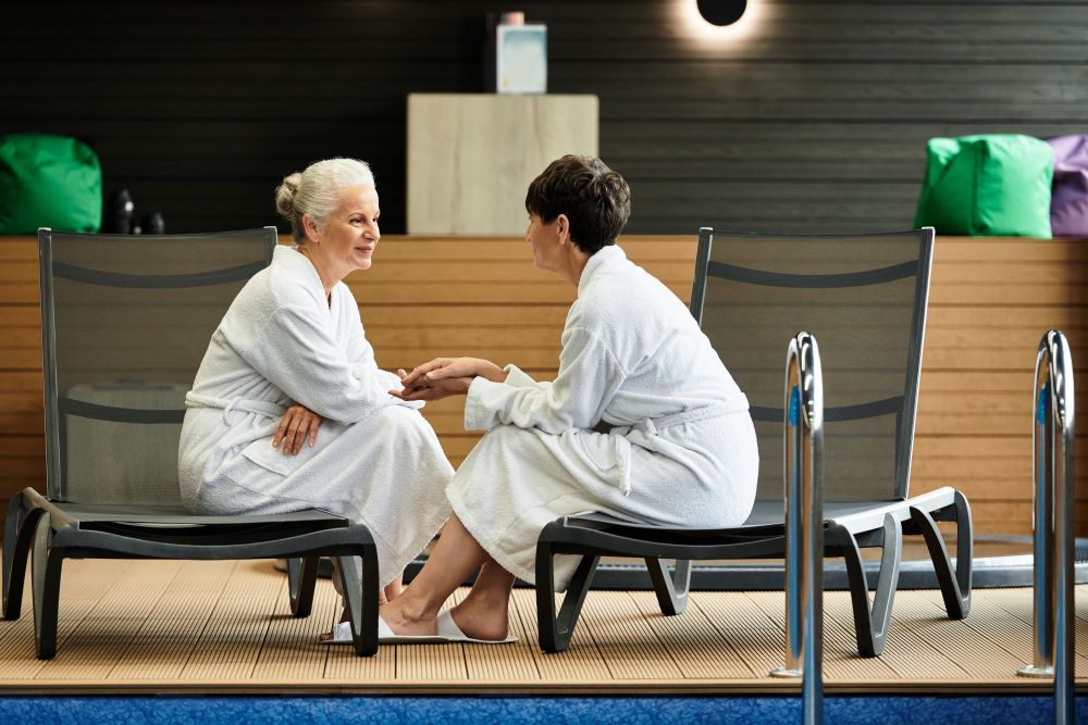 Two women in white robes sit on lounge chairs by a pool, engaged in a conversation and enjoying a spa-like atmosphere.