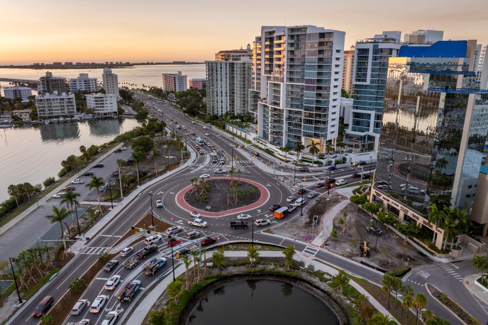 Traffic flows through a roundabout surrounded by high-rise buildings along the waterfront in Sarasota, Florida at sunset.