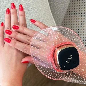 Hands with red nail polish using a FOREO hand care device, featuring a sleek design and glowing red lights in a cozy setting.