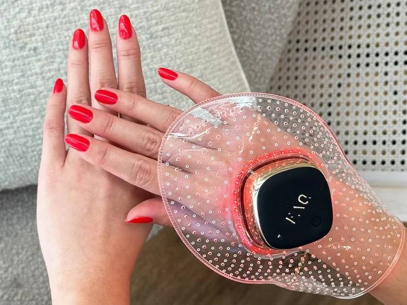 Hands with red nail polish using a FOREO hand care device, featuring a sleek design and glowing red lights in a cozy setting.